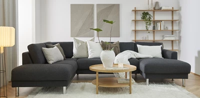 Stuemiljø med en mørk chaiselongsofa stående på hvidt gulvtæppe sammen med et sofabord, en træreol og billeder på væggen